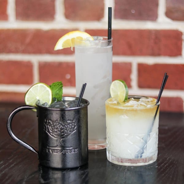 Three cocktails, one in a metal mug, one tall glass, and one short glass, against a brick wall.