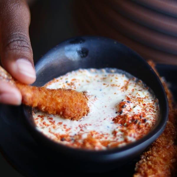 Hand dipping a crispy fried chicken strip into a creamy white sauce with red spices.