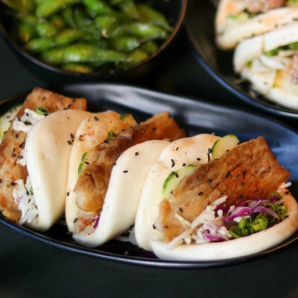A black plate of Boru Ramen's pork belly bao buns with cucumber, cabbage, and sesame seeds.
