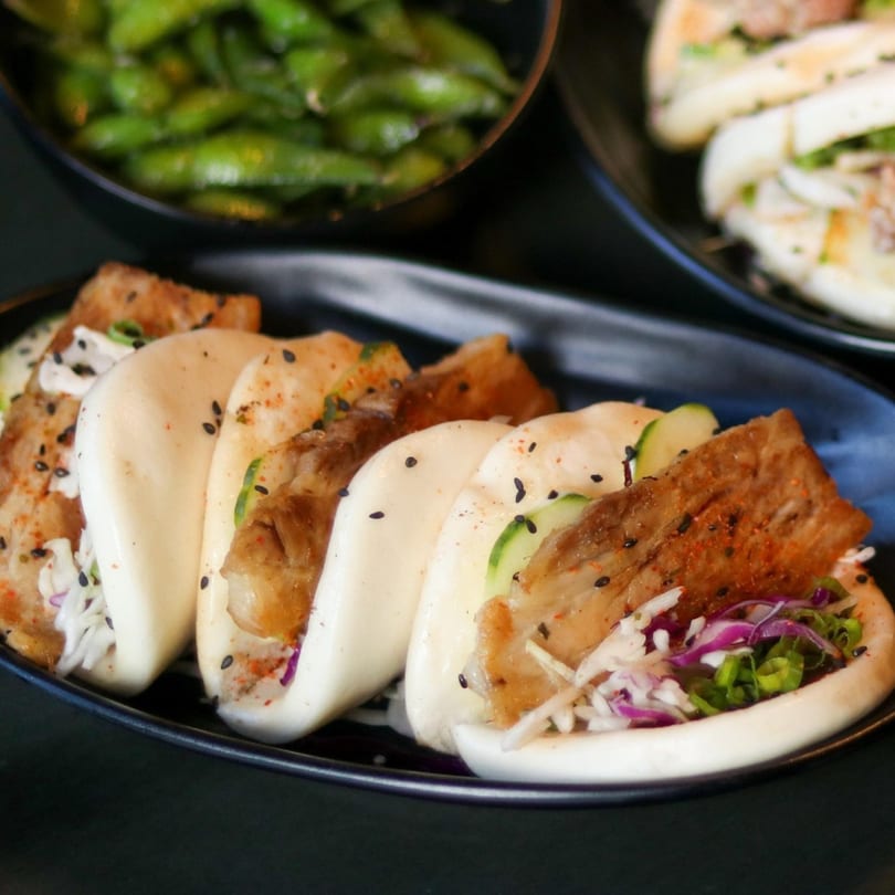 A black plate of Boru Ramen's pork belly bao buns with cucumber, cabbage, and sesame seeds.
