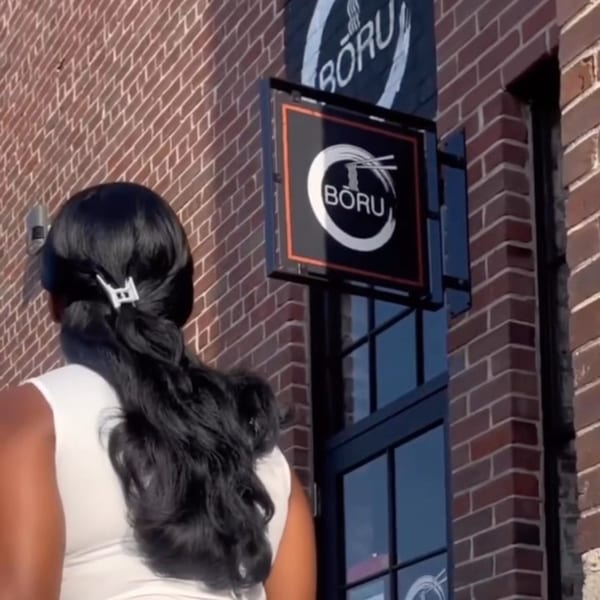 A person with dark hair and a white top stands near the Boru Ramen sign on a brick building.