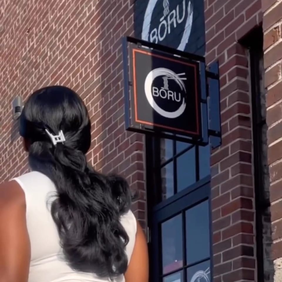 A person with dark hair and a white top stands near the Boru Ramen sign on a brick building.