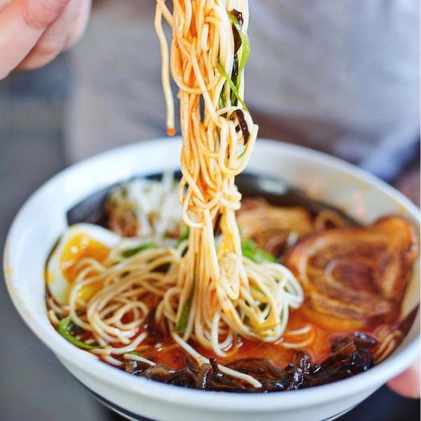 Chopsticks lifting a tangle of ramen noodles from a bowl with broth, pork, and a soft-boiled egg.