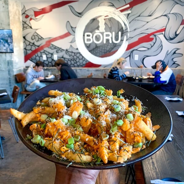 A hand holds a bowl of Boru Ramen's loaded fries with green onions, cheese, and seaweed.