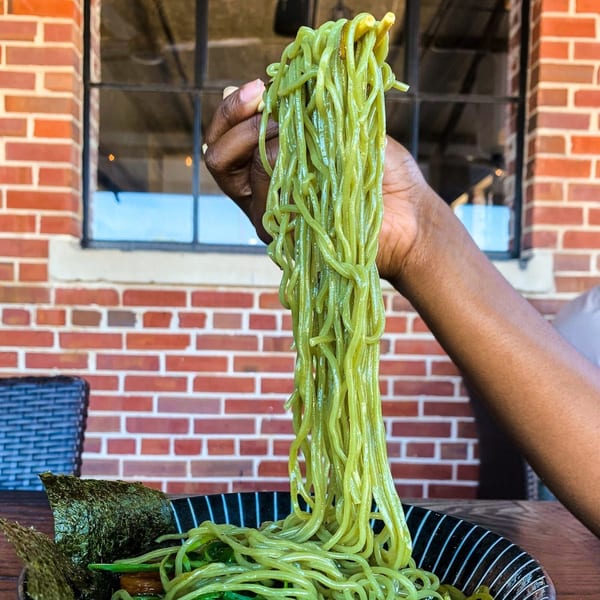 A hand holds up green ramen noodles with chopsticks from a black bowl with nori, against a brick wall.