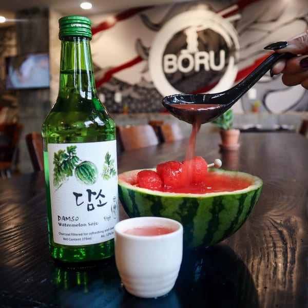 A bottle of Damso Watermelon Soju next to a watermelon bowl with a hand pouring more soju.