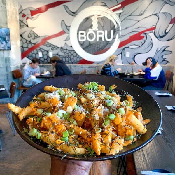 A close-up of Boru Ramen's loaded fries with green onions, against a restaurant interior.