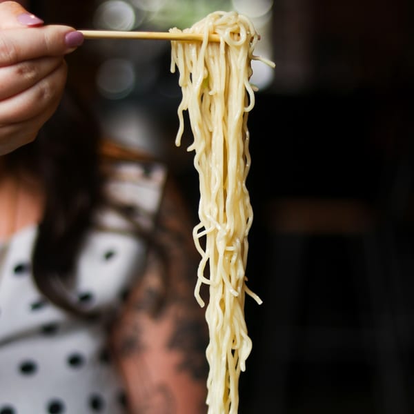 A hand holding chopsticks lifts a long strand of Boru Ramen noodles against a dark background.