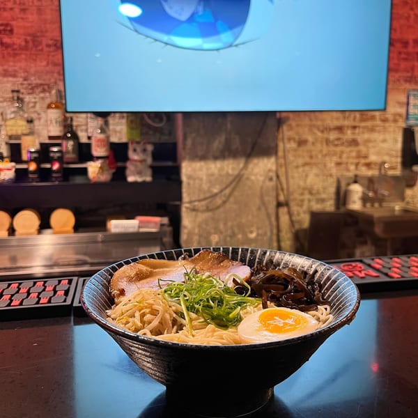 A bowl of Boru Ramen with pork, egg, and vegetables sits on a bar counter with a TV playing anime.