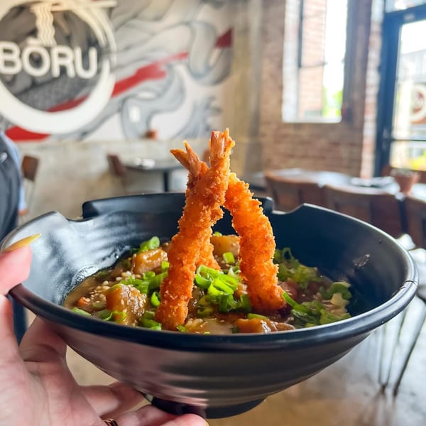 A person holds a black bowl of Boru Ramen's Japanese dish with fried shrimp and green onions.