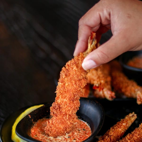 A hand dips a crispy fried shrimp tempura into a bowl of creamy sauce with red spices.