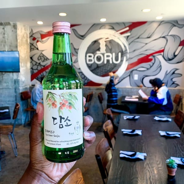 Hand holding a bottle of Damso Lychee Soju in Boru Ramen restaurant with a mural.