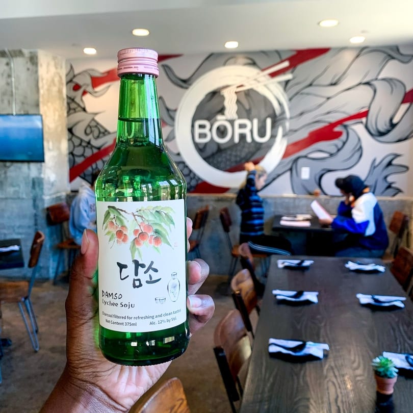 Hand holding a bottle of Damso Lychee Soju in Boru Ramen restaurant with a mural.